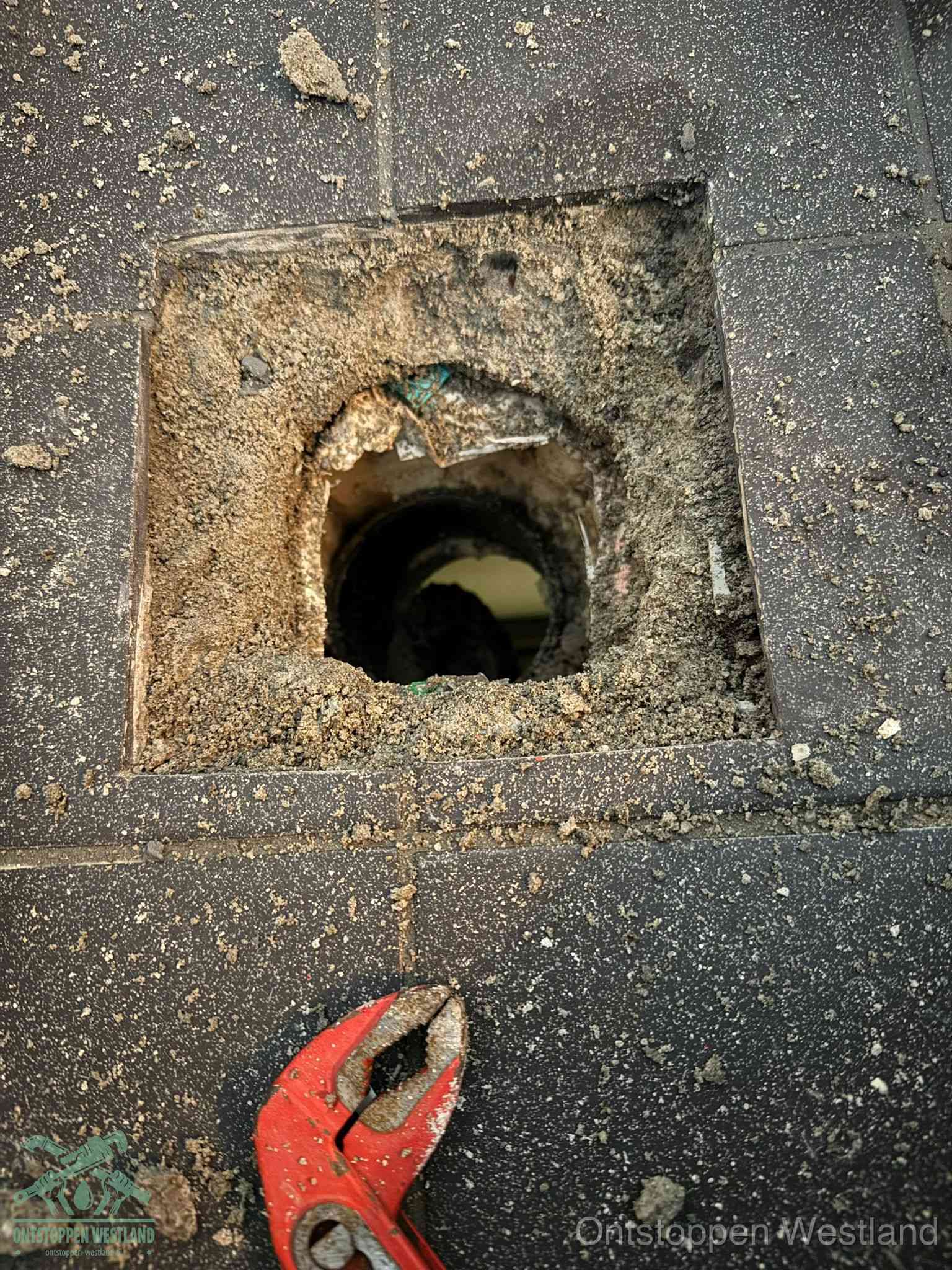 Een geopend rioolputje in een betontegelvloer met losgemaakt rooster en een zichtbare ronde afvoerbuis, het achterhalen waar de afvoerverstopping zit in Westland