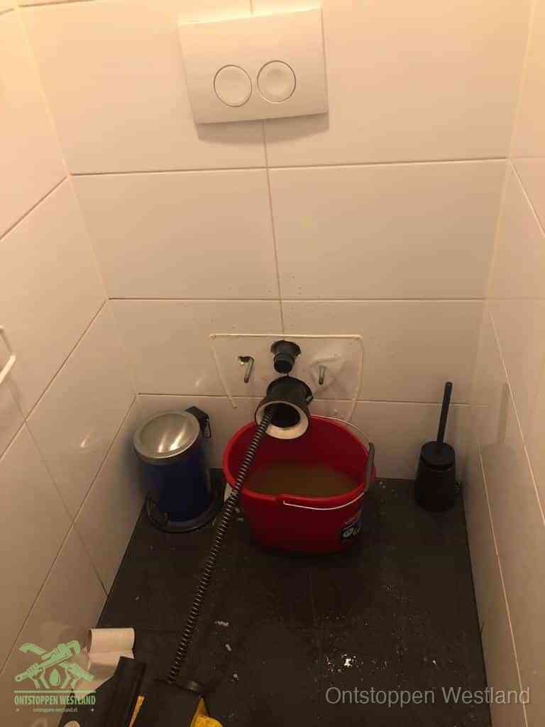 Een ontstopte wand-wc met verwijderde toiletpot waar een ontstoppingsveer in de wanduitgang verdwijnt, het achterhalen waar de verstopping zit in Westland