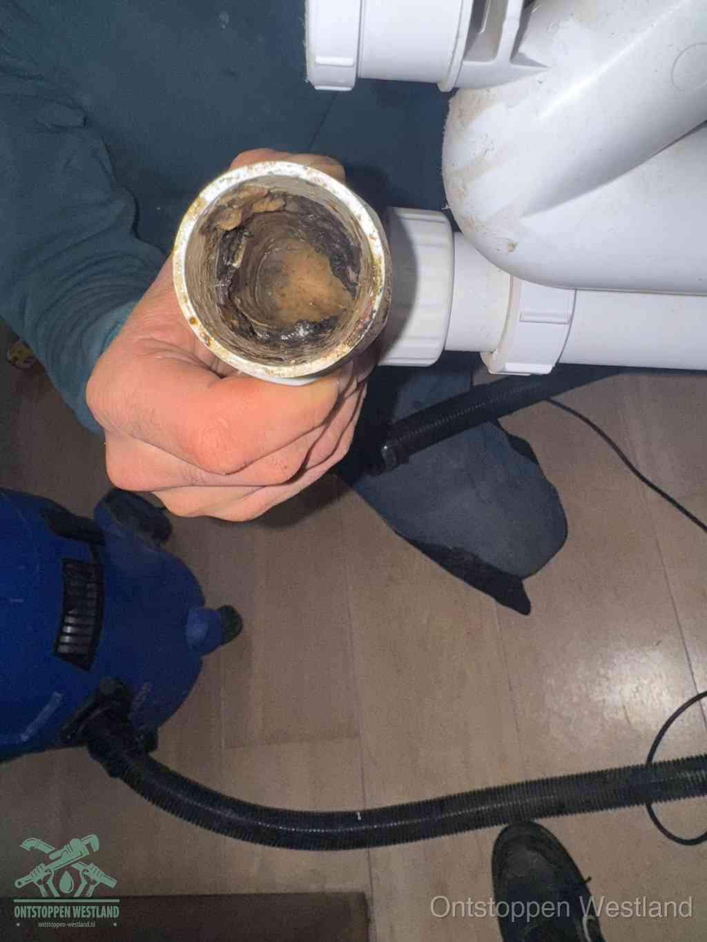 Een hand houdt een losgekoppeld PVC-afvoerbuisstuk vast dat van binnen volledig is dichtgeslibd met vet, vuil en aanslag, de meest voorkomende oorzaak van een verstopte afvoer in Westland