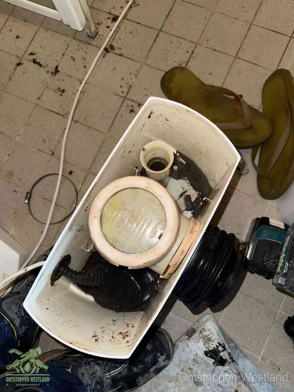 Een gedemonteerde toiletpot op de vloer met ontstoppingsveer en gereedschap ernaast, het verwijderen van een vastzittend voorwerp uit de wc in Westland