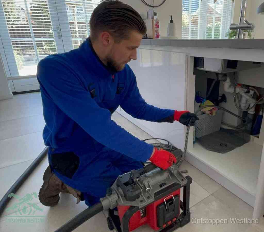 Een monteur met rode handschoenen knielt in een keuken en voert een elektrische ontstoppingsveer in tijdens het professioneel afvoer ontstoppen in Westland