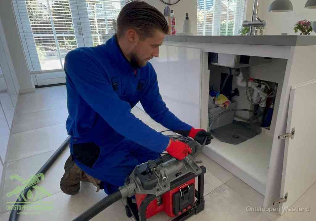 Een monteur voert een professionele elektrische ontstoppingsmachine in de afvoer onder de spoelbak tijdens het gootsteen ontstoppen in Westland